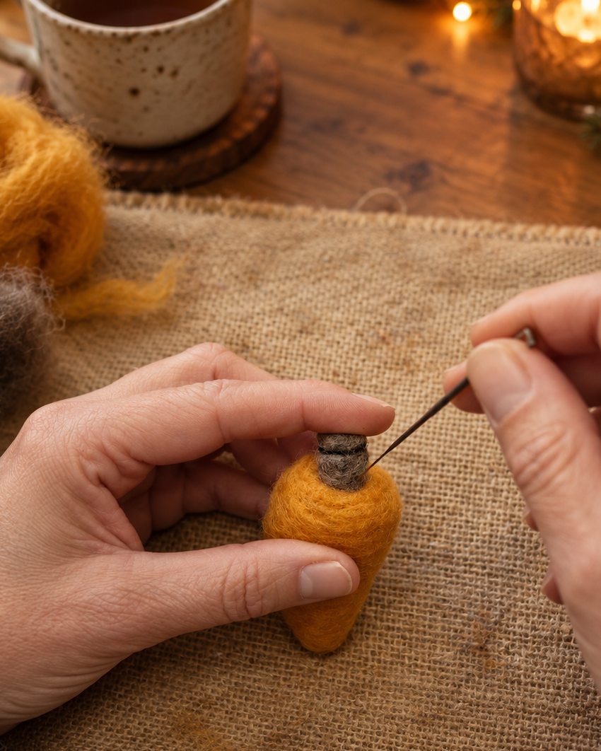 A close-up of hands crafting a small orange felted wool bottle with a wooden stopper using a needle tool, set on a burlap mat with a warm mug and candle in the background.
