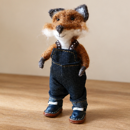 Needle felted fox wearing denim overalls and shoes on a wooden surface.